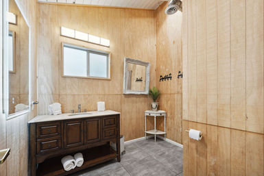 Cozy rustic wood-paneled bathroom with dark wooden vanity and marble countertop, frosted window, ornate mirror, small corner table with potted plant, gray tile floor and neatly rolled towels