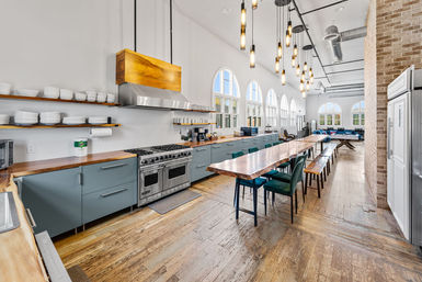 Sunlit communal industrial-style kitchen in a renovated loft with tall arched windows, exposed ductwork, hanging pendant lights, long live-edge wooden dining table with benches, teal lower cabinets, stainless double oven, and reclaimed hardwood floors.