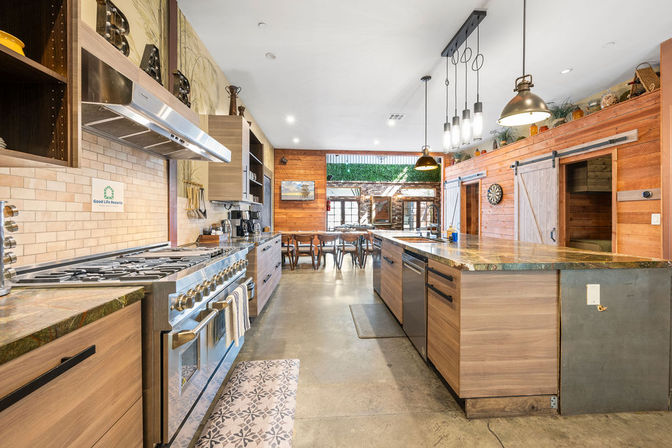 Spacious modern rustic open-concept kitchen with a large stone-topped island, stainless steel commercial range, wood-paneled walls and sliding barn doors, pendant lights, concrete floors, and a dining area opening to a sunny patio.