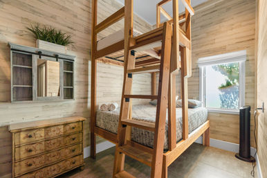 Cozy sunlit guest room with tall wooden bunk bed (twin-over-full), patterned bedding and rolled towels, rustic carved dresser, wall mirror with shelf and potted greenery, and a tower fan by the window.