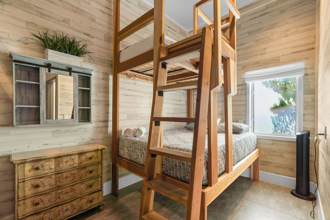 Cozy sunlit guest room with tall wooden bunk bed (twin-over-full), patterned bedding and rolled towels, rustic carved dresser, wall mirror with shelf and potted greenery, and a tower fan by the window.