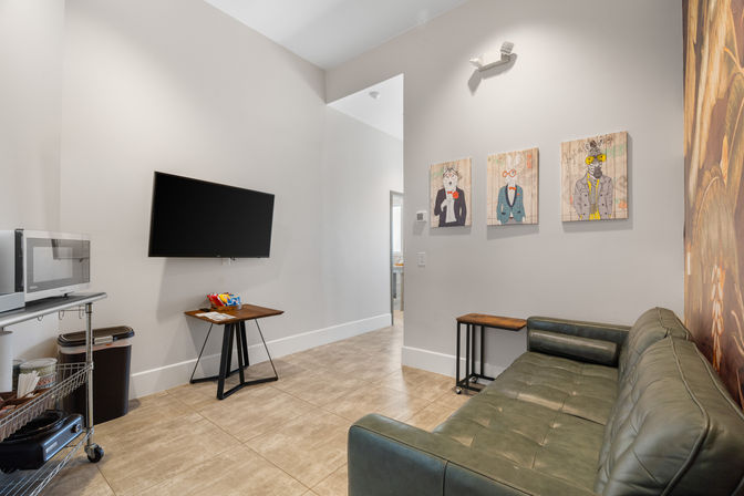 Modern lounge with wall-mounted flat-screen TV, green tufted leather sofa, small wooden tables, tile floor, microwave on a metal cart and three whimsical animal portraits on a light-gray wall.