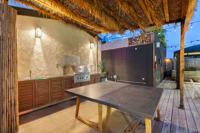Evening outdoor kitchen on a wooden deck with a tiki-style thatched roof, stainless gas grill and sink set in wood cabinetry, large concrete-topped island table, bamboo accents and string lights.