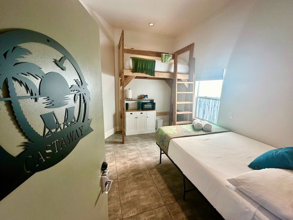 Sunny beach-cottage bedroom with wooden loft bunk and ladder, single bed with teal pillow, tiled floor, small kitchenette cabinet with microwave, window and neatly rolled towels.