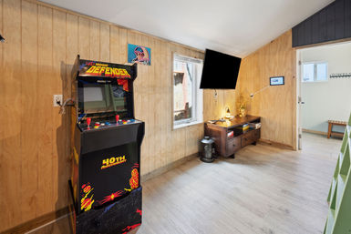 Retro home game room with a vintage Defender arcade cabinet, wall-mounted flat-screen TV, wood-paneled walls, light laminate floor, and a wooden console table with lamp and natural light from a window.