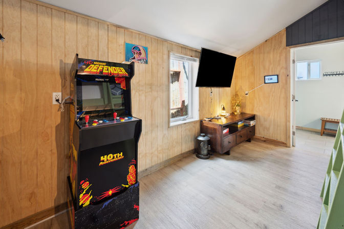 Retro home game room with a vintage Defender arcade cabinet, wall-mounted flat-screen TV, wood-paneled walls, light laminate floor, and a wooden console table with lamp and natural light from a window.