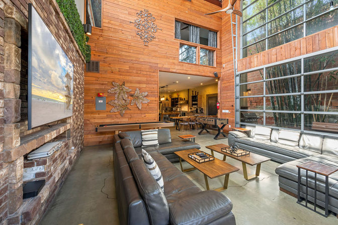 Sunlit modern industrial lounge in an urban loft with dark leather sectional sofas, wood-paneled walls, stone media wall with mounted TV, floor-to-ceiling glass garage door, coffee tables, foosball and shuffleboard, and an open kitchen dining area in the background.