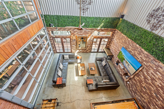 Loft-style living room with exposed brick walls and faux boxwood trim, large glass garage-door wall, concrete floor, two black leather sectional sofas around wooden coffee tables, wall-mounted TV showing a beach scene, ceiling fan, hanging industrial lights and a foosball table.