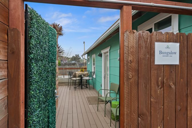 Private turquoise bungalow deck seen through a wooden gate, featuring a hedge panel, bistro table with chairs, wicker seating and palm trees in a fenced courtyard — cozy coastal-style outdoor dining area.