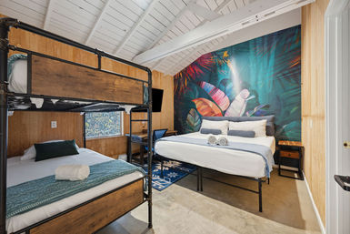 Vacation rental bedroom with metal-frame bunk beds and a queen bed in front of a colorful tropical leaf mural, white vaulted ceiling, wood-paneled walls, small desk and wall TV, concrete floor and rolled towels on the beds.