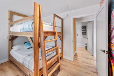 Contemporary bright bedroom with solid wood bunk beds, plaid bedding, light oak floors, white walls, and an open door to a hallway.