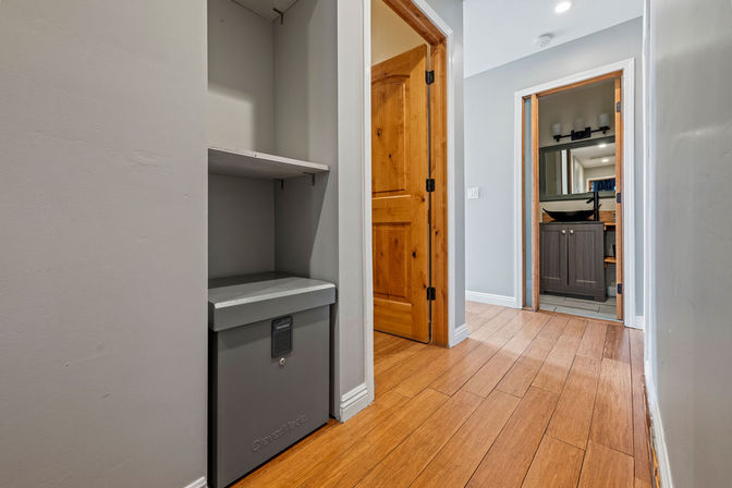 Interior hallway with warm hardwood floors, gray walls, built-in shelf and gray lockbox, open wooden door revealing a bathroom with dark vanity and mirror.