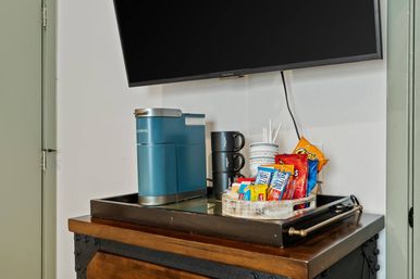 Mini coffee station in a hotel room: teal single-serve coffee maker, stacked black mugs and disposable cups with stirrers, tray of chips and snack bars on a wooden dresser under a wall-mounted TV.