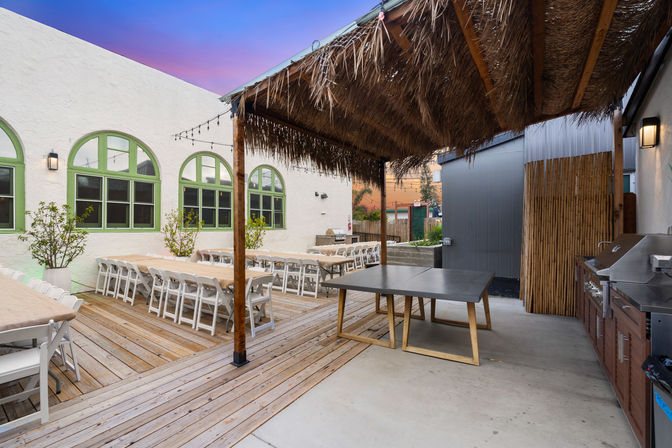 Twilight outdoor patio dining area on a wooden deck with long banquet tables and white folding chairs, green arched windows on a white stucco wall, a thatched palapa over a game table, string lights and a stainless-steel grill.