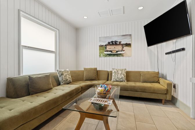 Cozy modern lounge with olive-green sectional, glass coffee table with snacks, wall-mounted TV, white shiplap walls and tiled floor