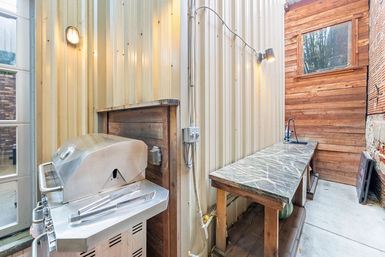 Narrow outdoor kitchen patio with stainless steel gas grill, long veined stone countertop and sink, wood-plank and corrugated metal walls, and wall-mounted lights.