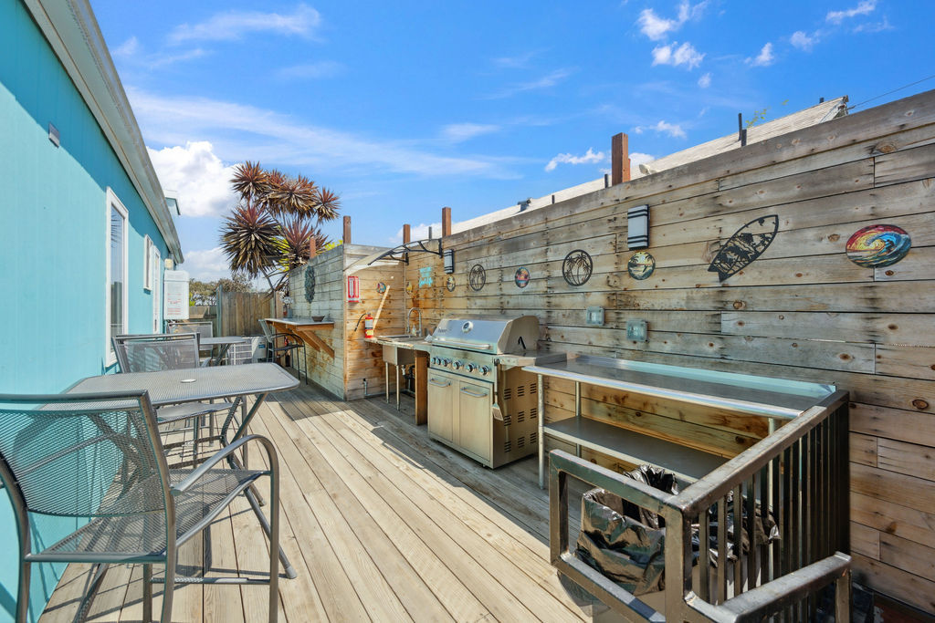 Sunny beachside wooden deck with stainless steel gas grill and prep counters, metal bar tables and chairs by a turquoise exterior, palm trees and ocean-themed wall decor under a bright blue sky.