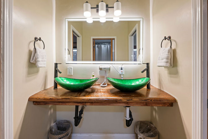 Bright bathroom double vanity with two green glass vessel sinks on a rustic wooden countertop, matte-black faucets, illuminated rectangular mirror, wall light and towel rings on neutral beige walls.