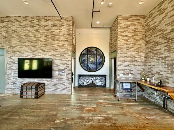 Sunlit industrial loft interior with exposed light brick walls, distressed wood plank floor, wall-mounted flat-screen TV above a vintage trunk, round stained-glass window over an ornate iron console, and a wooden counter with sink and metal shelving.