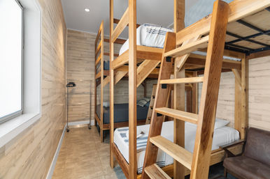 Bright hostel dorm room with multi-level wooden bunk beds and ladders, wood-paneled walls, tiled floor, natural light from a window, simple striped linens and a chair.