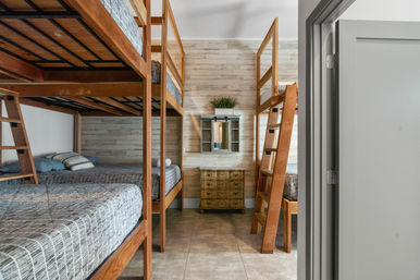 Cozy coastal-style bunk bedroom in a vacation rental with two wooden double-decker bunk beds, gray plaid bedding and ladders, weathered shiplap accent wall, small mirrored cabinet and distressed dresser on tiled floor.