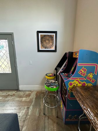 Corner of a home game room with a retro Ms. Pac-Man arcade cabinet, two colorful bar stools, framed abstract wall art, stained-glass front door, and worn wooden floor.