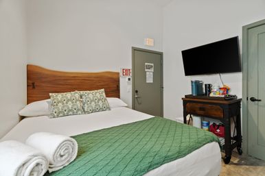 Cozy boutique hotel room with queen bed, wooden headboard, green quilt and patterned pillows, wall-mounted TV and a small snack and coffee station on a wooden cart by the door.