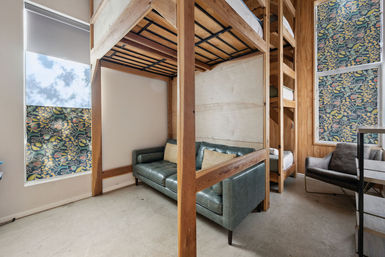Cozy compact loft-style room with wooden bunk beds, green leather sofa, floral-patterned window shades, gray armchair, and light concrete floor.