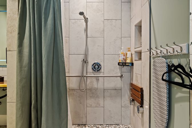 Accessible walk-in tiled shower with a green curtain, handheld showerhead, horizontal grab bar, fold-down wooden seat, pebble floor and toiletries on a corner shelf.