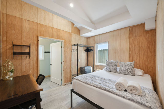 Cozy wood-paneled guest bedroom with white linens, patterned pillows, rolled towels on the bed, metal clothing rack, small desk and natural window light