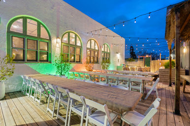 Cozy evening outdoor patio dining area on a wooden deck with long tables, white folding chairs, arched windows on a stucco wall, colorful uplighting and hanging string lights