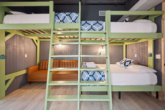 Cozy, group-friendly vacation rental bunk room with light-green lofted bunk beds, blue geometric bedding, gray wood-paneled walls and a tan leather sofa.