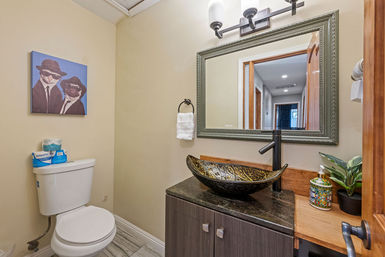 Small modern half-bath with beige walls, white toilet, quirky framed cat artwork, dark marble vanity topped with a decorative glass vessel sink, black faucet, framed mirror, potted plant and colorful soap dispenser.
