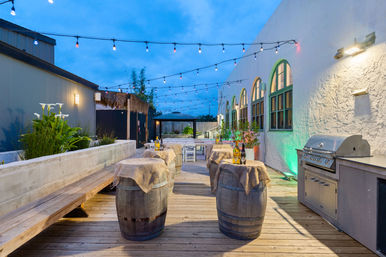 Evening rooftop patio deck with hanging string lights, wooden barrel cocktail tables draped in burlap topped with wine bottles and glasses, arched green‑trim windows, potted plants and a stainless gas grill.