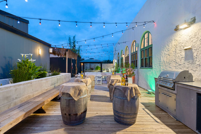 Evening rooftop patio deck with hanging string lights, wooden barrel cocktail tables draped in burlap topped with wine bottles and glasses, arched green‑trim windows, potted plants and a stainless gas grill.