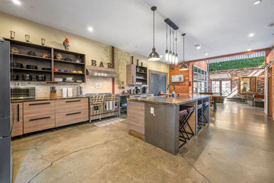 Industrial-chic open-concept urban loft kitchen with a large island and bar stools, stainless steel range, pendant lights, wood cabinetry, polished concrete floors and exposed brick leading to a sunlit seating area.