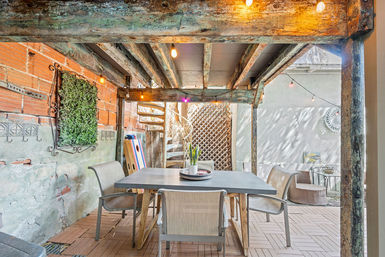 Cozy urban outdoor patio under a rustic wooden pergola with string lights, dining table and chairs, potted centerpiece, spiral metal staircase, and exposed brick and plaster walls.