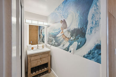 Compact coastal-style bathroom with a large surf wave mural featuring a surfer, rustic wood vanity with white countertop and lighted mirror