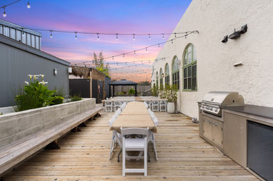 Rooftop wooden deck patio at sunset with a long communal table and white folding chairs, string lights overhead, stainless gas grill, arched windows, potted plants and bench seating for outdoor dining