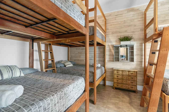 Cozy vacation-rental bunk room with sturdy wooden bunk beds, gray patterned bedding and ladders, rustic dresser, shiplap walls and potted plant — bright, group-friendly sleeping space.