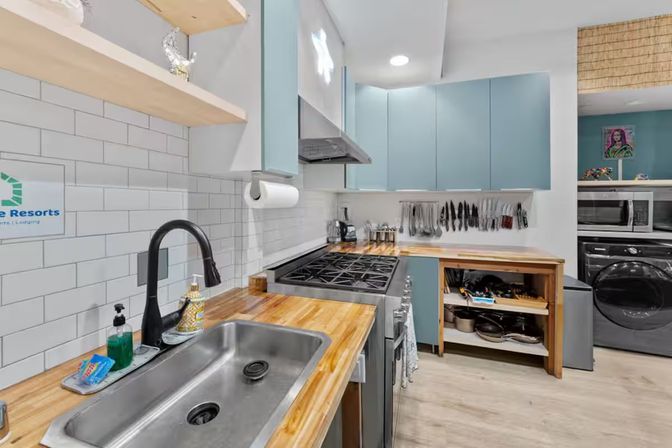 Bright modern kitchen with butcher-block countertops, subway tile backsplash, blue cabinets, stainless sink, gas range, hanging utensils and adjacent laundry nook