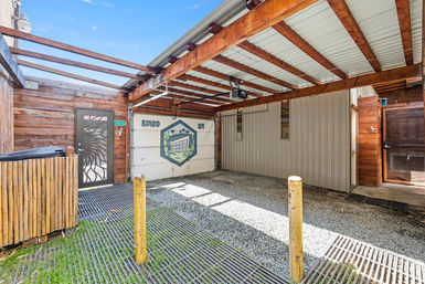 Sunlit covered carport with exposed wooden beams and corrugated metal roof, gravel floor, yellow bollards, wood-paneled walls and a decorative painted garage door mural