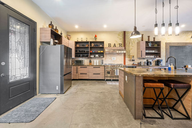Bright urban loft-style kitchen with concrete floor, stainless steel refrigerator and six-burner range under 'BAR' letters, wood cabinets with open shelving, stone-topped island with leather bar stools and industrial pendant lights.