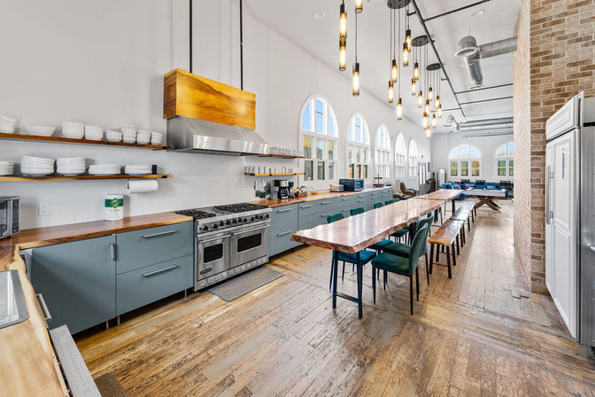 Bright industrial loft-style communal kitchen with a long live-edge wooden dining table, teal lower cabinets, stainless steel range and hood, hanging pendant lights, arched windows and reclaimed wood floors.