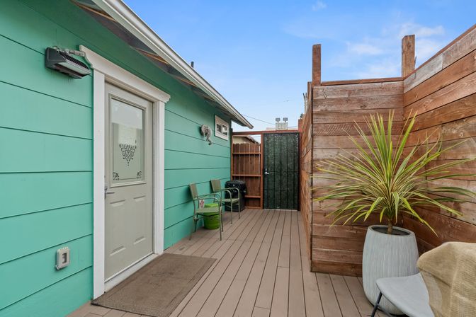 Cheerful turquoise cottage deck with white-trimmed door, wood privacy fence, potted spiky plant, two green chairs, doormat and a gated entry under blue sky