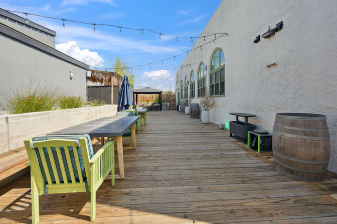 Sunlit rooftop wooden deck patio with string lights, long communal tables, lime-green chairs, barrels and arched green-framed windows under a bright blue sky