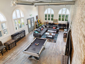 Industrial-style open loft game room with pool table, gray sectional sofa with colorful pillows, arcade cabinets, communal desks, large arched windows, exposed ductwork and hardwood floors