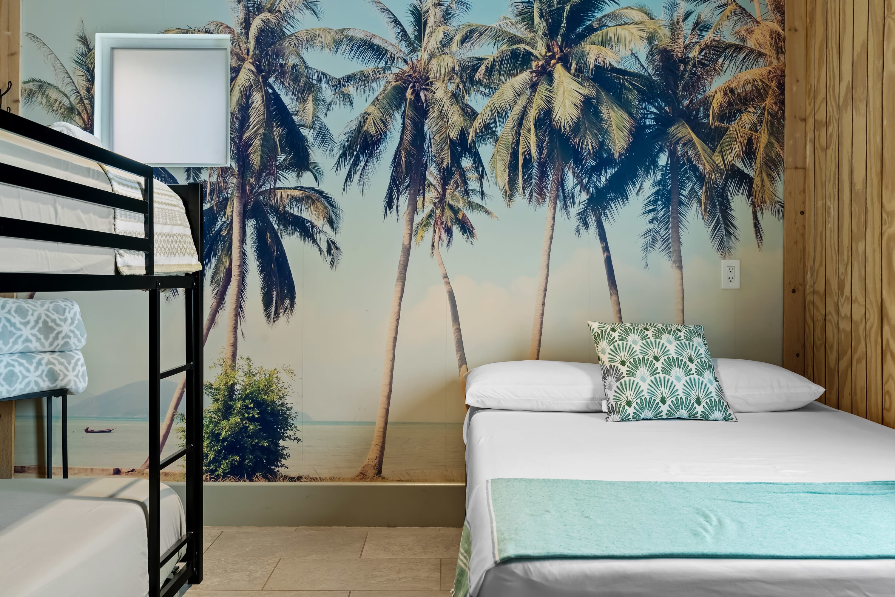 Beachy guest room with oversized palm-tree mural, black metal bunk bed on the left and a double bed on the right with a tropical-pattern pillow and teal throw — bright coastal décor.