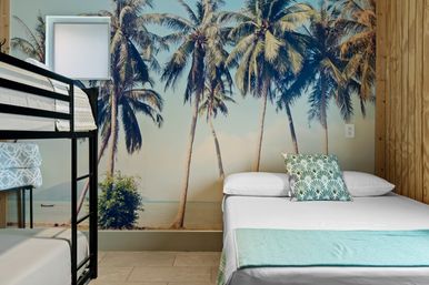Beachy guest room with oversized palm-tree mural, black metal bunk bed on the left and a double bed on the right with a tropical-pattern pillow and teal throw — bright coastal décor.