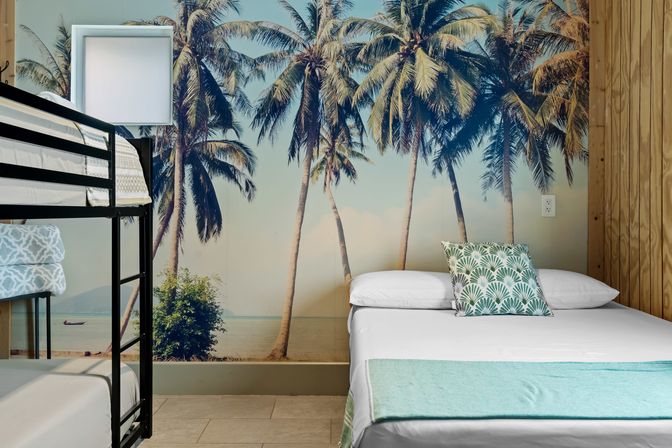 Beachy guest room with oversized palm-tree mural, black metal bunk bed on the left and a double bed on the right with a tropical-pattern pillow and teal throw — bright coastal décor.